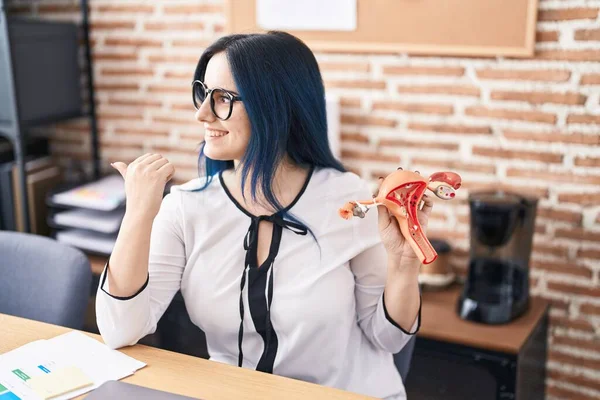Young girl with blue hair holding model of female genital organ at the office complaining for menstruation pain pointing thumb up to the side smiling happy with open mouth
