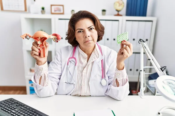 Middle age hispanic doctor woman holding anatomical female genital organ and birth control pills relaxed with serious expression on face. simple and natural looking at the camera.