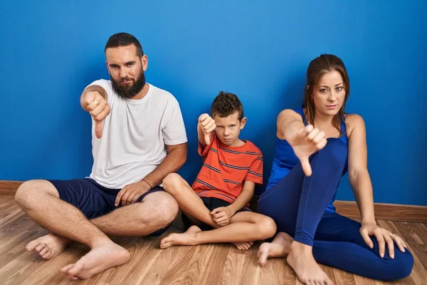 Family of three sitting on the floor at home looking unhappy and angry showing rejection and negative with thumbs down gesture. bad expression.