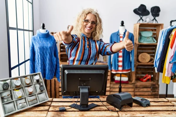 Middle age blonde woman working as manager at retail boutique approving doing positive gesture with hand, thumbs up smiling and happy for success. winner gesture.