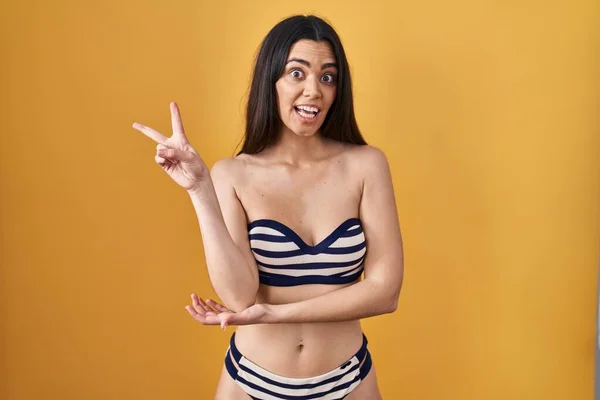 Young brunette woman wearing bikini over yellow background smiling with happy face winking at the camera doing victory sign with fingers. number two.