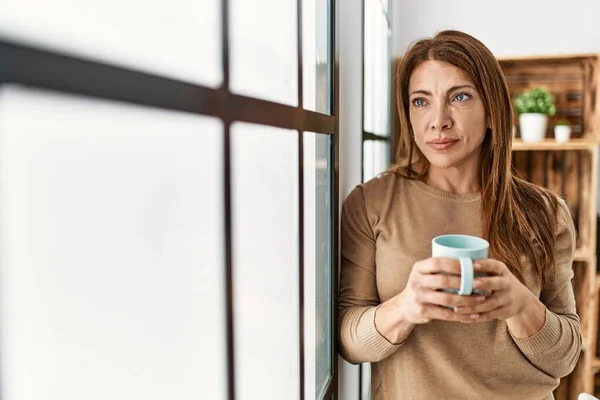 Middle age caucasian woman with serious expression drinking coffee standing at home.