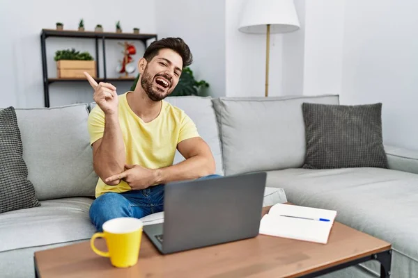 Young man with beard using laptop at home with a big smile on face, pointing with hand and finger to the side looking at the camera.