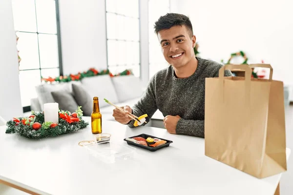 Young hispanic man eating sushi sitting by christmas tree at home