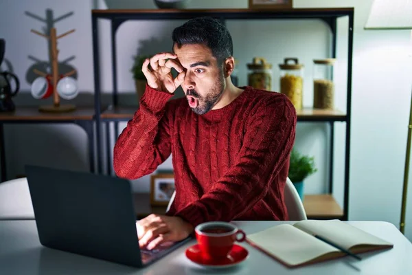 Young hispanic man with beard using computer laptop at night at home doing ok gesture shocked with surprised face, eye looking through fingers. unbelieving expression.