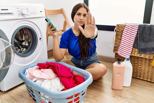Young brunette woman doing laundry using smartphone doing stop sing with palm of the hand. warning expression with negative and serious gesture on the face.