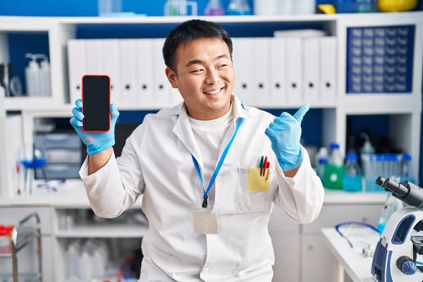 Young chinese man working at scientist laboratory holding smartphone pointing thumb up to the side smiling happy with open mouth