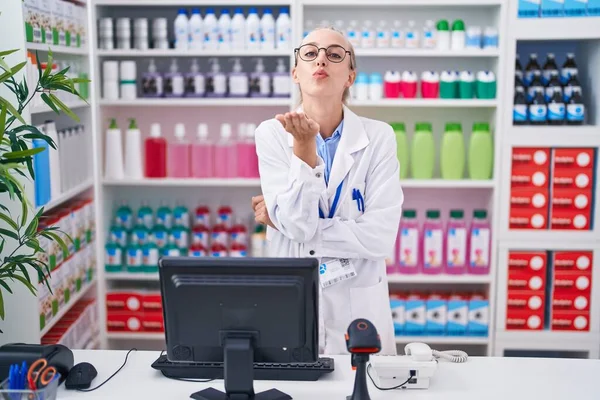 Young caucasian woman working at pharmacy drugstore looking at the camera blowing a kiss with hand on air being lovely and sexy. love expression.