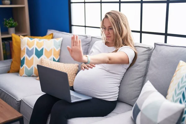 年轻的孕妇坐在家里的沙发上 用手提电脑 张开手 用严肃而自信的表情 做着手势 做着防守动作 — 图库照片
