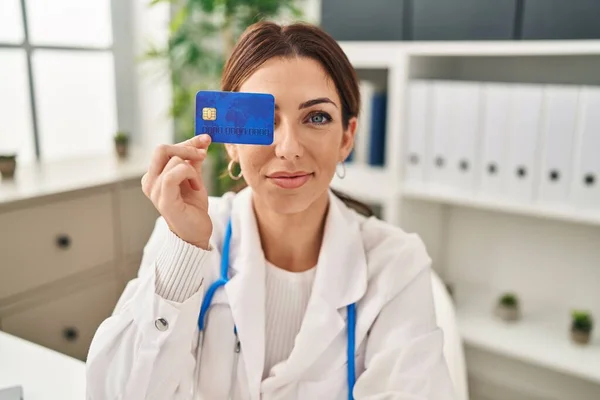 Young hispanic woman wearing doctor uniform holding credit card over eye at clinic
