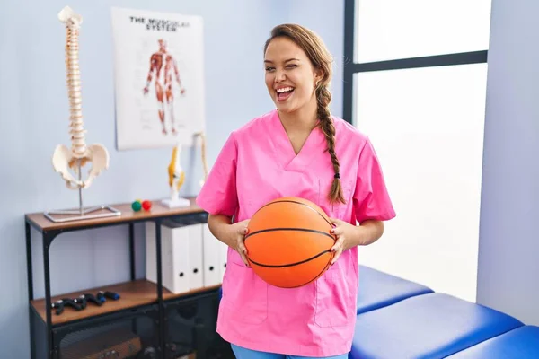 Young hispanic woman working at physiotherapy clinic holding basketball ball winking looking at the camera with sexy expression, cheerful and happy face.
