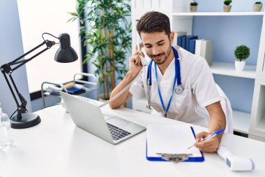 Young hispanic man wearing doctor uniform talking on the telephone working at clinic