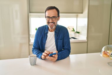 Orta yaşlı bir adam akıllı telefon kullanıp evde kahve içiyor.