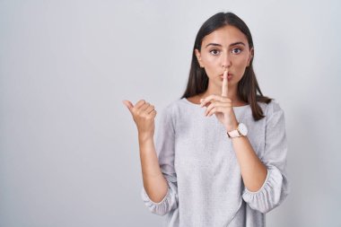 Young hispanic woman standing over white background asking to be quiet with finger on lips pointing with hand to the side. silence and secret concept. 