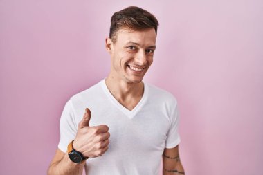 Caucasian man standing over pink background doing happy thumbs up gesture with hand. approving expression looking at the camera showing success. 