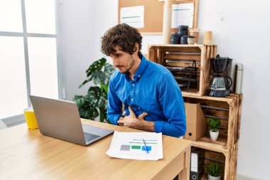 Young hispanic man with beard working at the office using computer laptop with hand on stomach because nausea, painful disease feeling unwell. ache concept. 