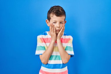 Mavi arka plan üzerinde duran genç beyaz çocuk yorgun eller yüzünü, depresyonu ve üzüntüyü örten, sorun yüzünden üzgün ve sinirli. 