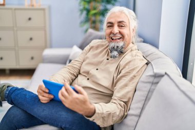 Middle age grey-haired man watching video on touchpad sitting on sofa at home