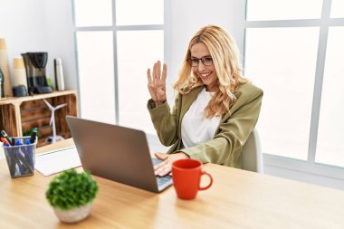 Beautiful blonde woman working at the office with laptop showing and pointing up with fingers number four while smiling confident and happy. 