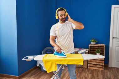 Young hispanic man listening to music ironing clothes at laundry room