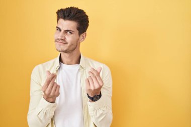 Sarı arka planda duran İspanyol genç adam elleriyle para hareketi yapıyor, maaş istiyor, milyonerlik işi yapıyor. 