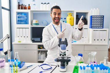 Sakallı, bilim adamı laboratuvarında çalışan, gülümseyen ve kameraya bakan, iki eli ve parmağı yanda olan genç İspanyol bir adam.. 