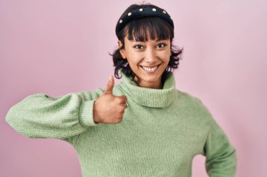 Young beautiful woman standing over pink background doing happy thumbs up gesture with hand. approving expression looking at the camera showing success. 