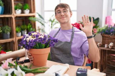 Caucasian blond man working at florist shop waiving saying hello happy and smiling, friendly welcome gesture 