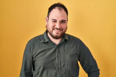 Plus size hispanic man with beard standing over yellow background winking looking at the camera with sexy expression, cheerful and happy face. 