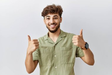 Young arab doctor man standing over isolated background success sign doing positive gesture with hand, thumbs up smiling and happy. cheerful expression and winner gesture. 