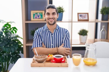 Hispanic man with long hair sitting on the table having breakfast happy face smiling with crossed arms looking at the camera. positive person. 