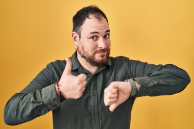 Plus size hispanic man with beard standing over yellow background doing thumbs up and down, disagreement and agreement expression. crazy conflict 