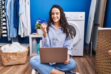 Çamaşır odasında çamaşır makinesi bekleyen bir İspanyol kadın dizüstü bilgisayar ve kredi kartı kullanıyor.