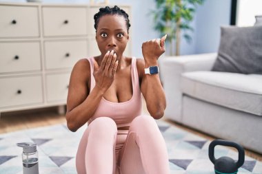 Beautiful black woman using smart watch training at home covering mouth with hand, shocked and afraid for mistake. surprised expression 