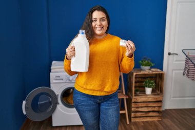 Young hispanic woman holding detergent bottle smiling with a happy and cool smile on face. showing teeth. 