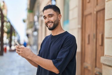 Genç Arap adam sokakta akıllı telefon kullanarak kendinden emin gülümsüyor.