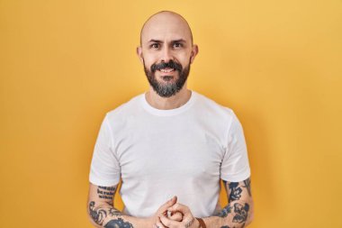 Sarı arka planda dikilen dövmeleri olan genç İspanyol adam ellerini kavuşturup gülümseyerek gevşemiş ve neşeliydi. Başarı ve iyimserlik 