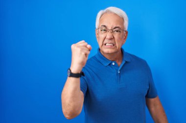 Mavi arka planda duran gri saçlı, sinirli ve öfkeli bir orta yaşlı adam öfkeyle bağırırken yumruğunu kaldırıyor. öfke ve agresif konsept. 