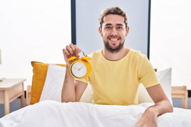 Hispanic man with beard holding alarm clock in the bed looking positive and happy standing and smiling with a confident smile showing teeth 