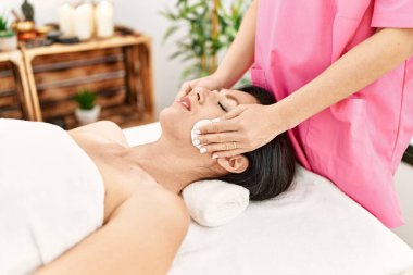Middle age hispanic woman having facial treatment cleaning face at beauty center