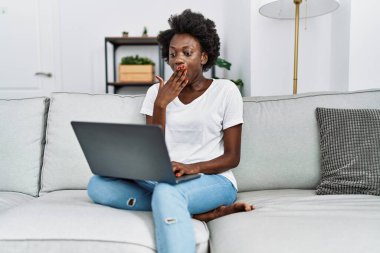 African young woman using laptop at home covering mouth with hand, shocked and afraid for mistake. surprised expression 