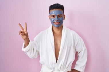 Young hispanic man wearing beauty face mask and bath robe smiling looking to the camera showing fingers doing victory sign. number two. 