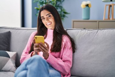 Evdeki kanepede oturan, akıllı telefon kullanan genç İspanyol kadın.