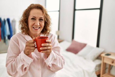 Orta yaşlı beyaz kadın yatak odasında kahve içiyor..