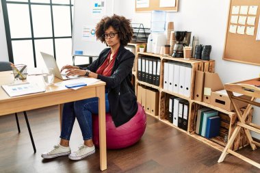 Genç Afro-Amerikalı kadın, topun üzerinde oturmuş ofiste çalışıyor.