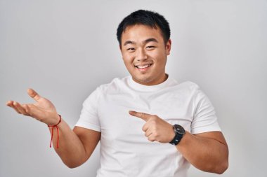 Young chinese man standing over white background amazed and smiling to the camera while presenting with hand and pointing with finger. 