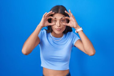 Brunette young woman standing over blue background trying to open eyes with fingers, sleepy and tired for morning fatigue 