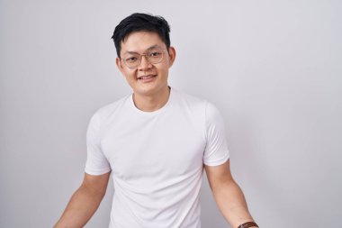 Young asian man standing over white background smiling cheerful with open arms as friendly welcome, positive and confident greetings 