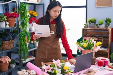 Çiçekçide dizüstü bilgisayar kullanarak kitap okuyan Çinli bir kadın.
