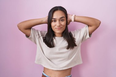 Genç İspanyol kadın pembe arka planda duruyor rahatlıyor ve esniyor, kolları ve elleri başının arkasında mutlu bir şekilde gülümsüyor. 